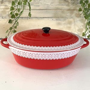 May include: An oval red enameled cast iron Dutch oven with a matching lid and two side handles. The exterior of the pot features a decorative white lace trim band encircling the upper rim, and the lid is topped with a small black knob.