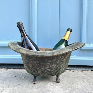 May include: An antique bronze-colored metal champagne bucket with ornate floral detailing, holding two bottles of champagne. The bucket has a bathtub shape with claw feet and is set against a blue background.