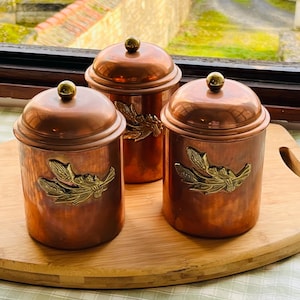 Op de afbeelding: Drie koperen potten met messing details en deksels. Elke pot heeft een gouden knop en een messing bladontwerp. De potten worden gepresenteerd op een houten dienblad.