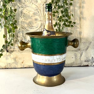 Op de afbeelding: Decoratieve champagnekoeler met messing accenten, groene, witte en blauwe emaille en twee handvatten. Er staat een champagnefles in. De emmer heeft een brede rand en een voet.