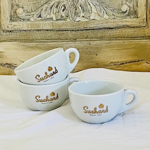 May include: White ceramic Suchard coffee cups stacked on a white surface. Each cup has a gold logo and the words "Suchard depuis 1826". The cups have a rounded shape with a handle.