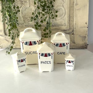 Op de afbeelding: Een set van vijf crèmekleurige keramische keukencanners met deksels. Elke kan heeft een handvat en is versierd met een rood, wit en blauw ontwerp. De kannen zijn gelabeld met "SUCRE", "CAFE", "PATES", "POIVRE" en "EPICES".