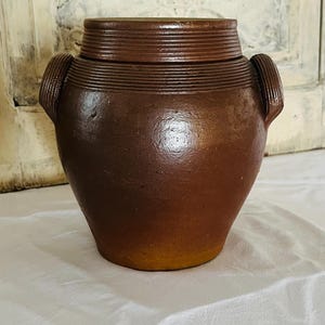 Könnte beinhalten: Ein brauner Keramiktopf mit Deckel. Der Topf hat einen abgerundeten Körper mit zwei kleinen Griffen und einem strukturierten Deckel. Der Topf ist etwa 20 cm hoch und 18 cm breit. Der Topf hat eine warme braune Farbe mit einem leichten Farbverlauf.