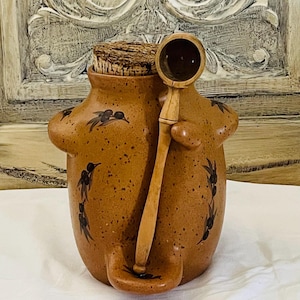 Puede incluir: Un tarro de cerámica marrón con tapón de corcho y cuchara de madera. El tarro tiene forma redondeada con dos asas laterales y un pequeño pico. Diseños florales negros decoran la superficie del tarro. La cuchara de madera descansa en el lateral.