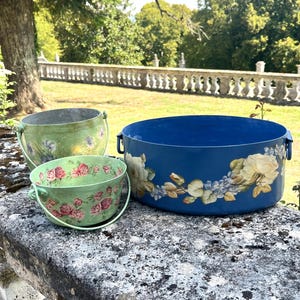 May include: Three metal containers including two small green buckets with handles and floral patterns, and one large blue circular basin with side handles featuring a painted design of white roses and blue flowers along the exterior side.