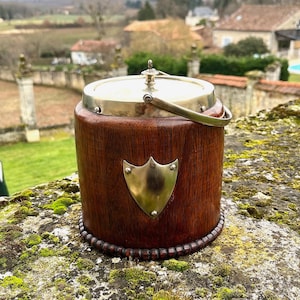 May include: A vintage wooden ice bucket with a brass lid and handle. The bucket features a brass shield emblem. The wooden body is a dark brown. The bucket is on a moss-covered stone, with a rural landscape in the background.