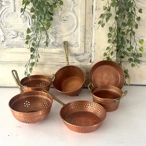 Op de afbeelding: Een set van zes miniatuur koperen keukengerei, waaronder potten, pannen en een vergiet, elk met een gehamerde textuur en messing handgrepen. De set bevat verschillende vormen en maten, allemaal met een warme, metalen afwerking.