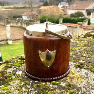 May include: A wooden ice bucket with a metal handle and lid. The bucket has a shield-shaped emblem on the front. The bucket is sitting on a stone wall covered in moss, with a blurred background of a landscape.