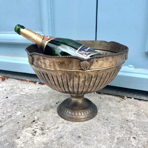 Op de afbeelding: Een antieke champagnekoeler in bronskleur met een gegroefd ontwerp en een voetstuk. Er staat een groene champagnefles in. De koeler heeft een geschulpte rand en staat op een betonnen oppervlak.