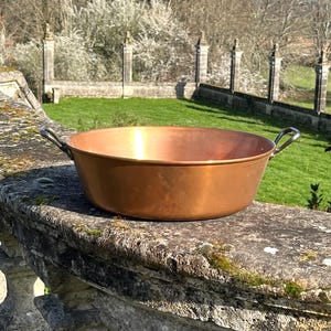 May include: A large copper bowl with two curved handles. The bowl has a wide, shallow design and a polished surface. It sits on a stone wall, with a blurred background of green grass and trees. The bowl is likely around 40 cm in diameter.