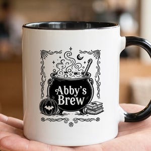 Puede incluir: Una taza de cerámica blanca con asa y borde interior negros. El frente presenta una ilustración gráfica negra de un caldero burbujeante, una calabaza con un murciélago y una pila de libros, con el texto Abby's Brew centrado en el diseño.