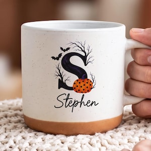 Puede incluir: Una taza de cerámica blanca con acabado moteado y base de color terracota. El frente presenta una letra S negra grande decorada con ramas de árboles desnudas, murciélagos volando y una calabaza naranja con lunares, con el nombre Stephen debajo.