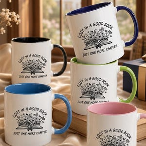 May include: Five white ceramic mugs with colored interiors and handles. Each mug features a black graphic of an open book with flowers and butterflies, with the text "LOST IN A GOOD BOOK" and "JUST ONE MORE CHAPTER" printed on the front.