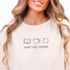 以下が含まれることがあります： 「read · sip · repeat」のテキストと、本、マグカップ、リサイクルのアイコンがプリントされたクリーム色のTシャツ。 ゆったりとしたフィット感のシャツです。 人は格子模様の白いマグカップを持っています。