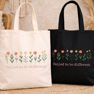 May include: Two tote bags, one cream and one black, with floral designs. Each bag has a row of colorful flowers and the text "Called to be different" printed in a light pink color.