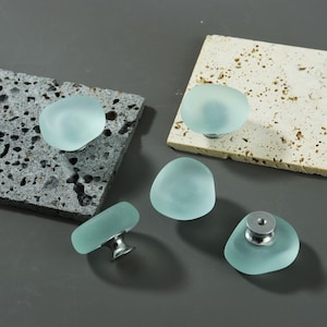 May include: A collection of five sea glass cabinet knobs in various orientations. The knobs are a soft, translucent blue-green color, with a smooth, rounded shape. Each knob is mounted on a silver-toned metal base.
