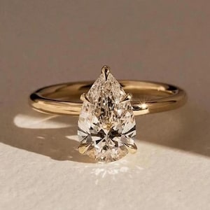 May include: A gold ring with a pear-shaped diamond. The diamond is the focal point, with a clear, multifaceted appearance. The band is a simple, polished gold. The ring is set against a neutral background, highlighting its elegance.