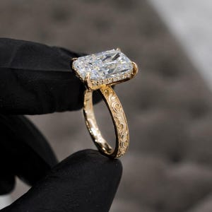 May include: A gold ring featuring a large rectangular radiant-cut clear gemstone set in a prong setting. The band is crafted from yellow gold and displays an intricate, embossed floral scrollwork pattern along the entire outer surface.