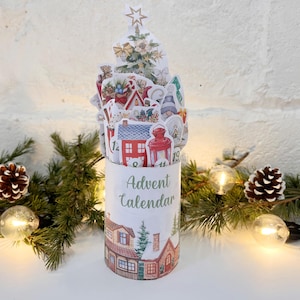 Könnte beinhalten: Ein zylindrischer Adventskalender mit einem Weihnachtsbaum-Design oben. Der Kalender zeigt Illustrationen von Häusern, Zwergen und einer roten Laterne. Die Worte "Adventskalender" sind auf der Seite aufgedruckt. Die Szene ist mit Tannenzapfen, Grün und Lichterketten dekoriert.
