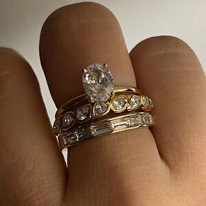 May include: A hand wearing three gold rings. The top ring features an oval-cut diamond. The middle ring has small round diamonds. The bottom ring has rectangular diamonds. The rings are set against a neutral background.