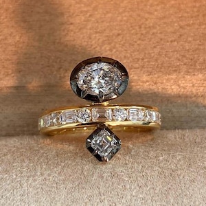 Puede incluir: Un anillo de oro con un diseño único, con un diamante de talla ovalada en la parte superior, una banda con diamantes más pequeños y un diamante de talla cuadrada en la parte inferior. El anillo se presenta sobre un fondo neutro.