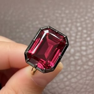 May include: A rectangular, ruby-red gemstone ring with a dark gray bezel. The ring's band is gold-colored. The gemstone is held in place by a dark gray, geometric setting. The ring is a statement piece.