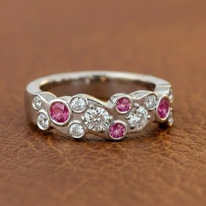 Peut inclure: Une bague en argent avec un motif ondulé, sertie d'une combinaison de pierres précieuses roses rondes et de pierres précieuses rondes claires. La bague est présentée sur une surface marron.