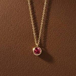 May include: A gold necklace featuring a small, round, red gemstone pendant. The delicate chain hangs against a brown backdrop. The pendant is set in a gold bezel.