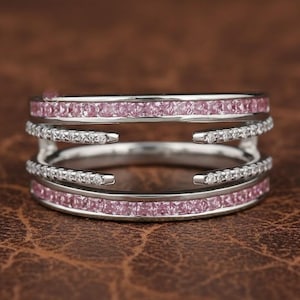 Può includere: Un anello d'argento con tre fasce. Le fasce superiore e inferiore sono decorate con piccole pietre preziose rosa. La fascia centrale è ornata da piccole pietre preziose trasparenti. L'anello è posto su uno sfondo di pelle marrone.