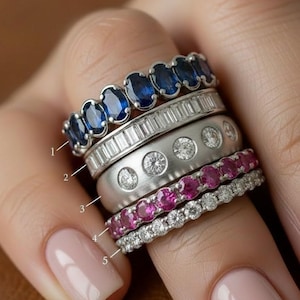 Puede incluir: Una colección de cinco anillos, cada uno con un diseño único. El anillo superior presenta piedras preciosas azules ovaladas, seguido de un anillo con piedras claras rectangulares. El tercer anillo es plateado con pequeñas piedras claras, y el cuarto tiene piedras preciosas rosas. El anillo inferior tiene piedras claras.