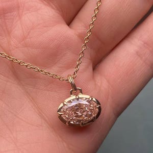 May include: A gold necklace with an oval pendant, featuring a light pink gemstone. The pendant is set in gold and suspended from a fine gold chain. The jewellery is shown against a neutral backdrop.