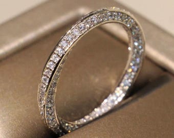 May include: A silver ring with a double band design, encrusted with numerous small, sparkling clear stones. The ring is set against a neutral, warm-toned background, highlighting its brilliance.