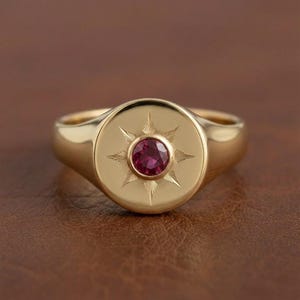 Peut inclure: Bague en or avec une face circulaire ornée d'un motif en forme d'éclat d'étoile et d'une pierre précieuse rouge ronde au centre. La bague est posée sur un fond marron, mettant en valeur l'or poli et la couleur de la pierre.