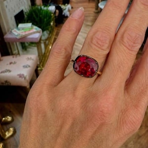 Pode incluir: Um anel de ouro com uma grande pedra preciosa vermelho-escura, lapidada em formato de almofada e engastada num bisel de metal escuro. A pedra tem um tom carmesim saturado com facetas internas visíveis que captam a luz.