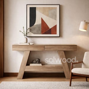 May include: A light-colored wooden console table with two drawers and a lower shelf. A framed abstract art piece hangs above the table. A small sculpture and books are on the shelf. The table is in a room with a rug and chair.