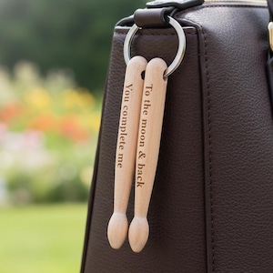 Puede incluir: Un bolso de cuero marrón oscuro con un llavero plateado que sostiene dos llaveros de madera con forma de baquetas. Los llaveros están grabados con las frases "You complete me" y "To the moon & back". El fondo es un jardín borroso.