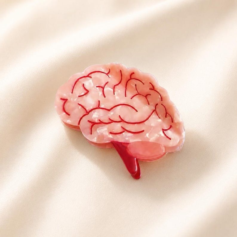 May include: A hair clip shaped like a human brain, featuring a translucent, pearlescent light pink base with intricate, deep red vein-like lines etched across the surface, and a solid, glossy dark red stem extending from the bottom section.