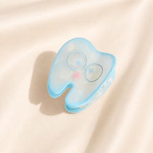 May include: A light blue hair claw clip shaped like a tooth with a translucent finish. The clip features two circular outlines for eyes and a small pink dot for a mouth on the front surface, with a metal spring mechanism visible on the side.