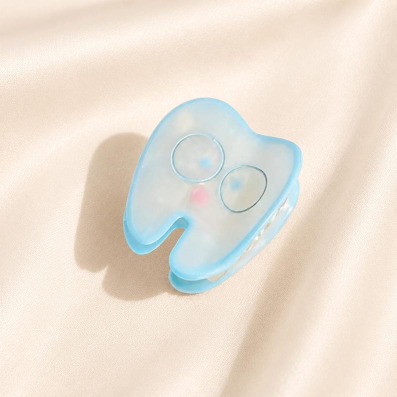 May include: A light blue hair claw clip shaped like a tooth with a translucent finish. The clip features two circular outlines for eyes and a small pink dot for a mouth on the front surface, with a metal spring mechanism visible on the side.