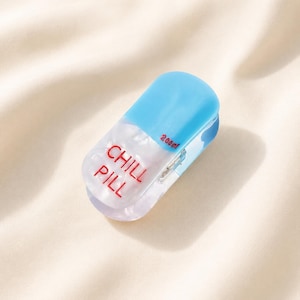 May include: A pill-shaped container with a glossy light blue top and a translucent white bottom. The text CHILL PILL is printed in red on the front, with 200ml printed in red on the side. The object has a smooth, rounded, and polished finish.