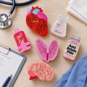 May include: A collection of acrylic medical-themed pins including a red heart labeled Jenny, a clear 1000ml flask labeled Dana, a pink syringe, a pink lung, a pink pill bottle labeled Muddy Anti Delulu Pills, and a pink brain labeled Lola.