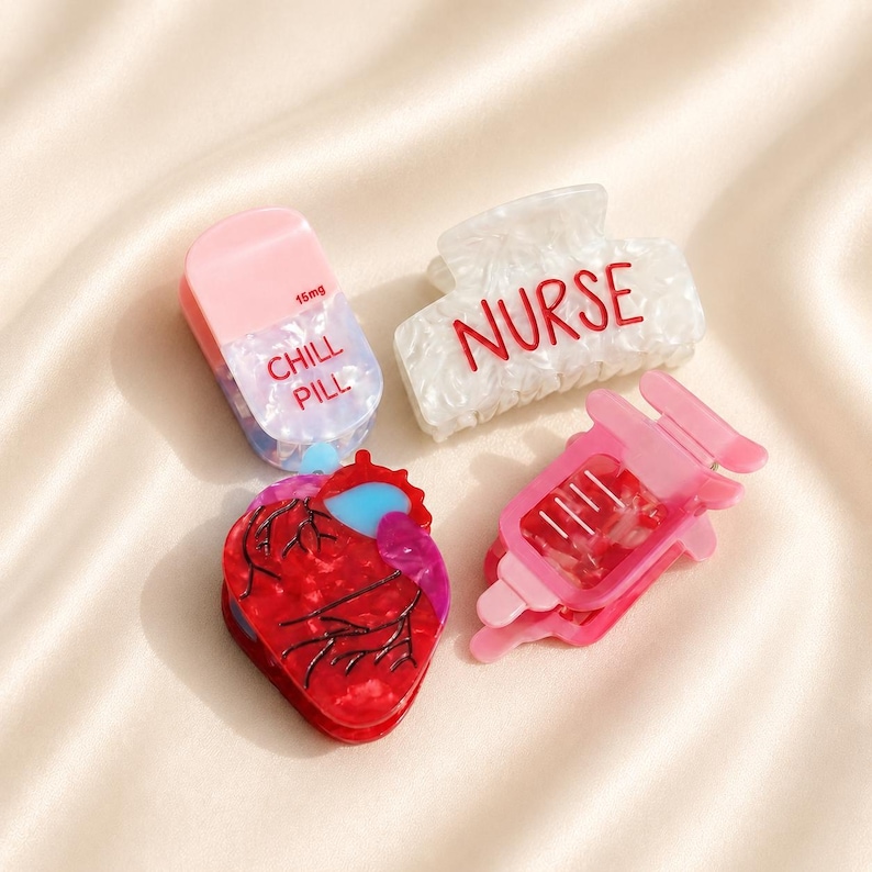 May include: Four hair clips featuring medical and health themes: a pink and white pill-shaped clip labeled CHILL PILL 15mg, a white marbled clip with the red text NURSE, a red anatomical heart clip with blue accents, and a pink syringe-shaped clip.