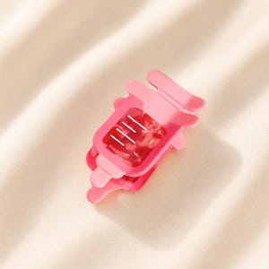 May include: A bright pink hair claw clip featuring a rectangular frame with rounded edges and a translucent, marbled texture. The clip has a spring-loaded mechanism with four visible silver metal teeth on the interior for securing hair.