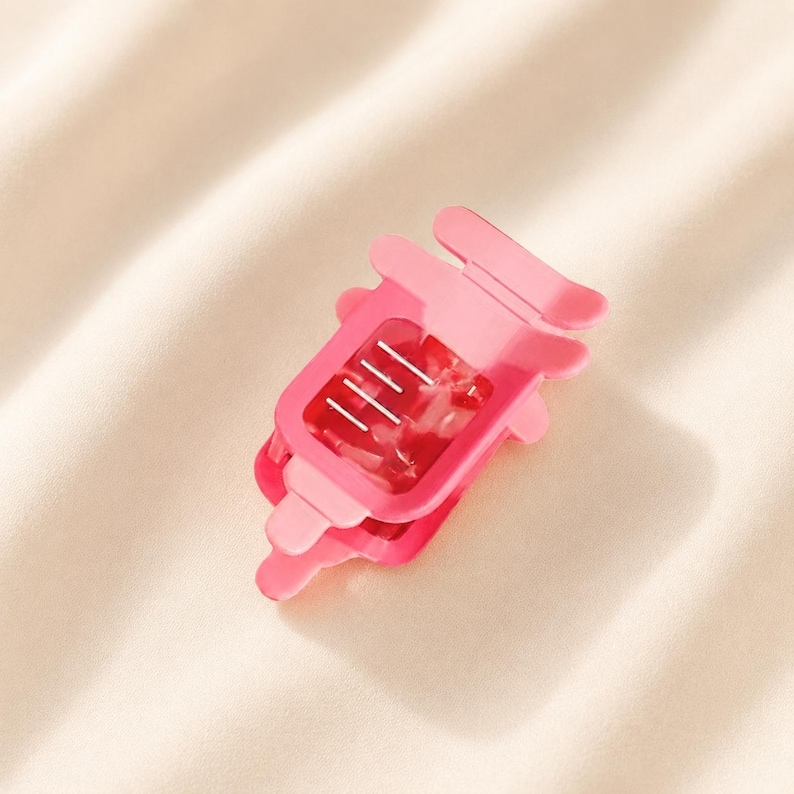 May include: A bright pink hair claw clip featuring a rectangular frame with rounded edges and a translucent, marbled texture. The clip has a spring-loaded mechanism with four visible silver metal teeth on the interior for securing hair.