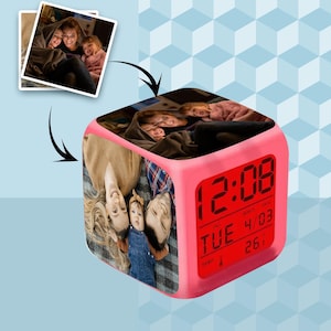Puede incluir: Un reloj despertador digital rosa con un collage de fotos familiares en sus lados. El reloj muestra la hora, la fecha y la temperatura en rojo. El reloj, con forma de cubo, está sobre una superficie azul claro. La hora es 12:08.