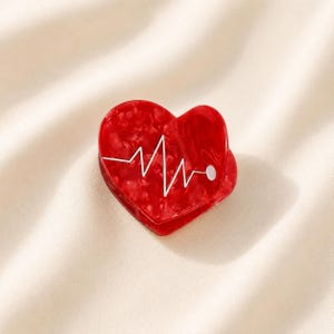 May include: A heart-shaped hair claw clip in a glossy red marbled finish. The surface features a white graphic line representing an electrocardiogram heartbeat rhythm, ending in a small white dot on the right side of the heart shape.