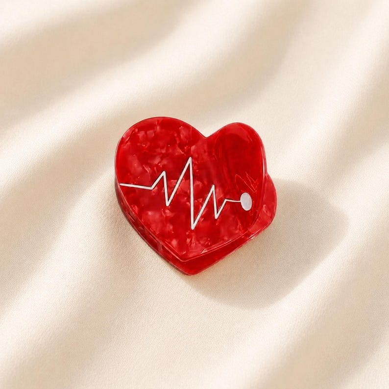 May include: A heart-shaped hair claw clip in a glossy red marbled finish. The surface features a white graphic line representing an electrocardiogram heartbeat rhythm, ending in a small white dot on the right side of the heart shape.