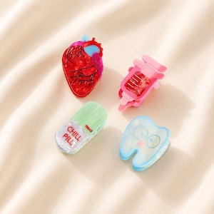 May include: Four hair clips featuring medical-themed designs: a red anatomical heart, a pink syringe, a light green pill capsule labeled CHILL PILL 200MG, and a light blue tooth with a face. Each clip is made of a smooth, glossy material.