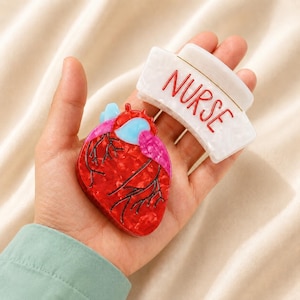 May include: A white hair claw clip with the word NURSE written in red letters and a separate anatomical heart hair claw clip featuring a marbled red, pink, and light blue design with black vein details, both made of polished acrylic material.