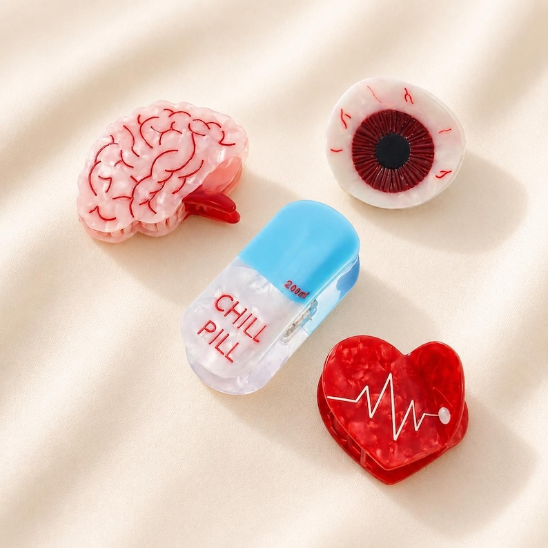 May include: Four hair clips featuring anatomical and novelty designs: a pink brain, a realistic human eyeball, a blue and white pill capsule labeled CHILL PILL with 200mg text, and a red heart with a white EKG heartbeat line graphic printed on it.